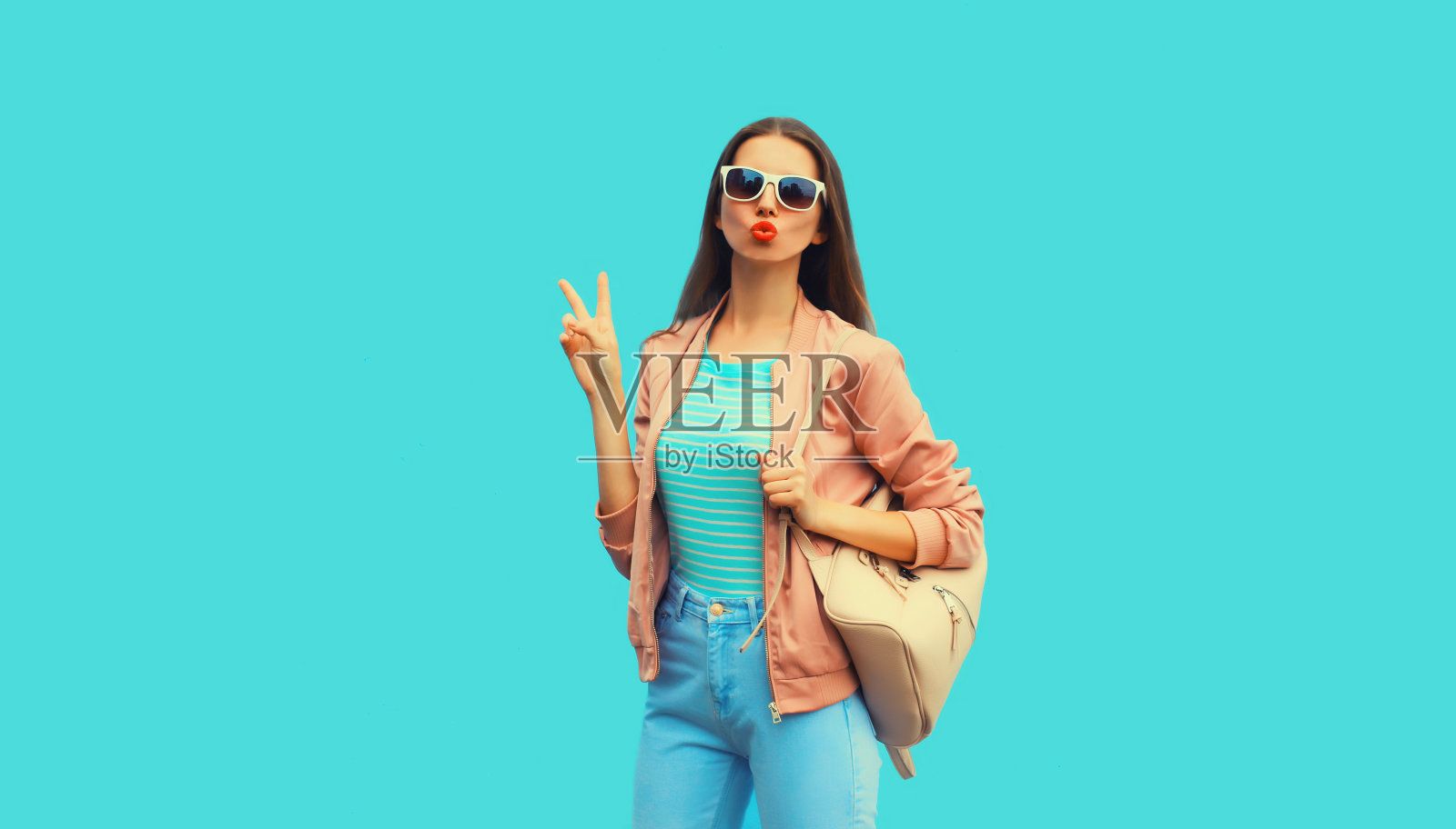 年轻女子的肖像摆造型吹她的嘴唇送空气吻背着背包,太阳镜在蓝色的背景照片摄影图片