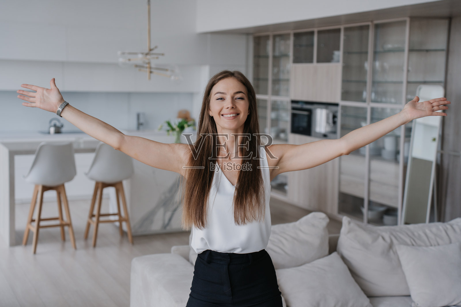 开朗的白人年轻女子随意摊开双手站在客厅欢迎拥抱。满意的意大利女孩对新公寓感到高兴,庆祝拥有。房地产经纪人介绍公寓照片摄影图片
