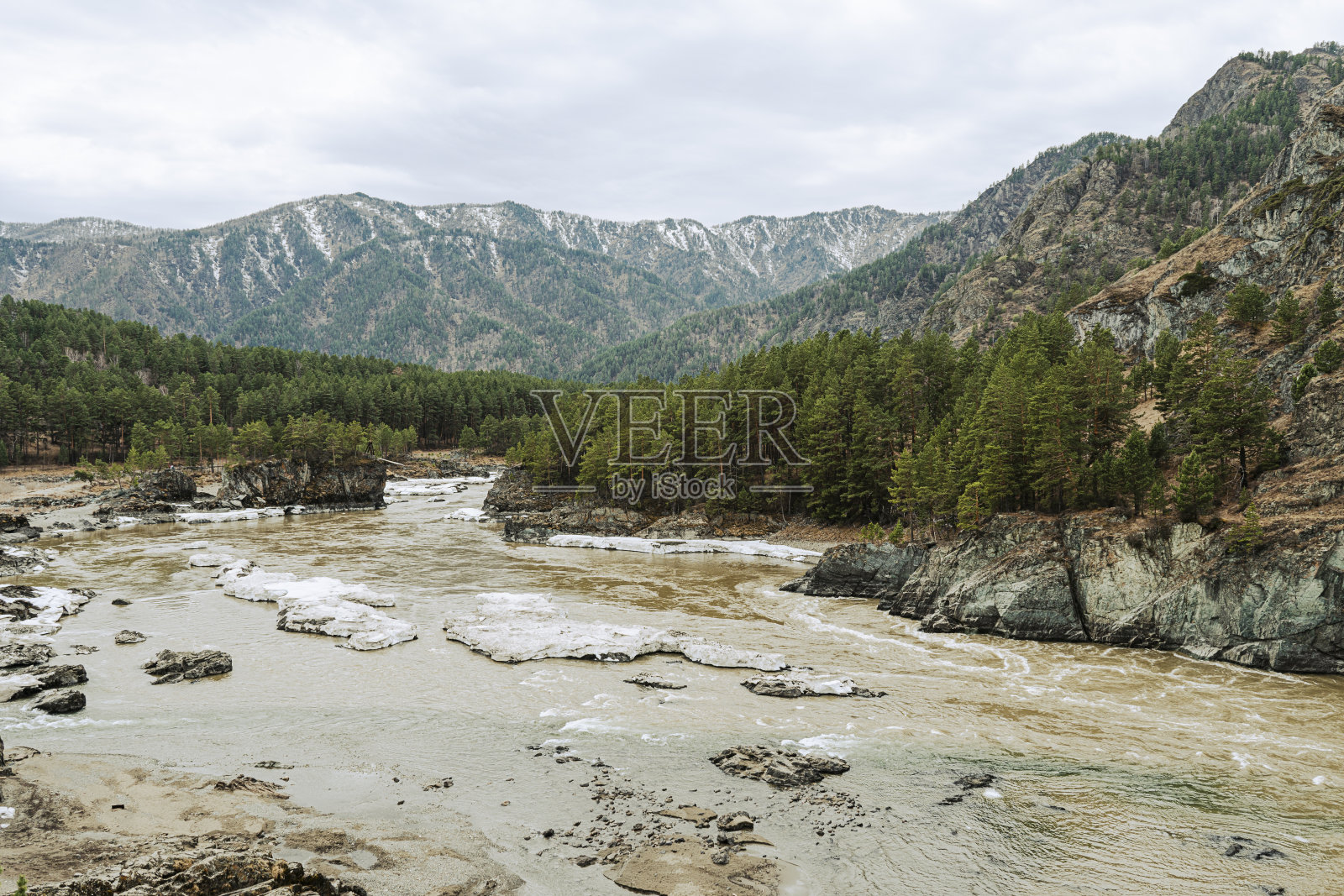 这条河在白雪覆盖的海岸之间流动。石头上结了一层蓬松的霜。雪堆中的乔木和灌木。群山映着蓝天。早春。照片摄影图片