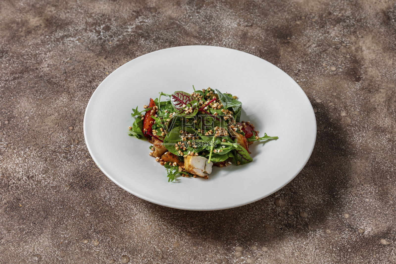 鹌鹑,辣椒粉,南瓜,什锦生菜,香蒜酱和荞麦谷物的热沙拉。食物放在一个圆圆的、白色的陶瓷深盘子里。盘子放在咖啡色的深色织物背景上。照片摄影图片