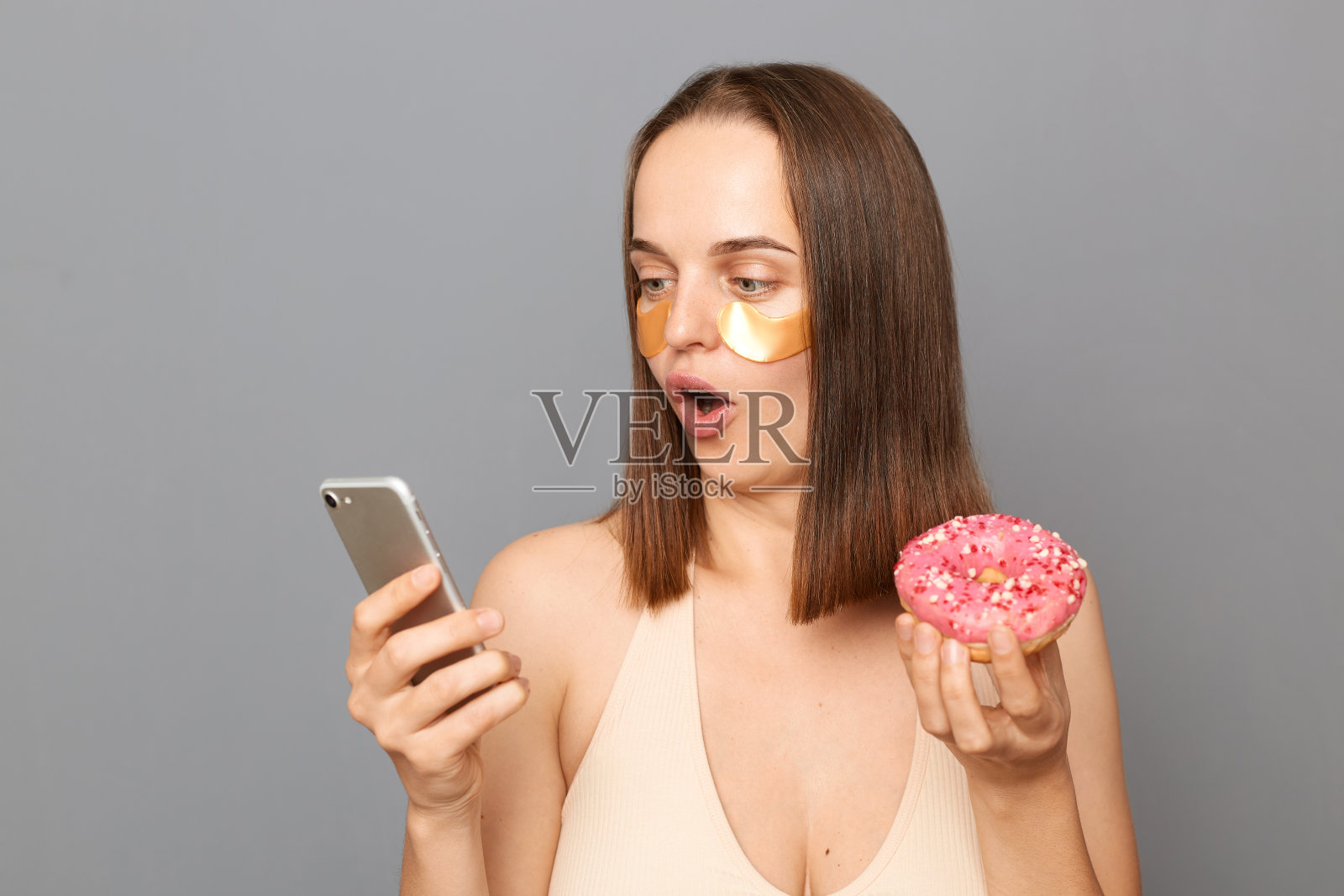 惊讶的年轻女子拿着手机和美味的甜甜圈蛋糕孤立在灰色背景,吃糖果和使用手机,检查社交网络。照片摄影图片