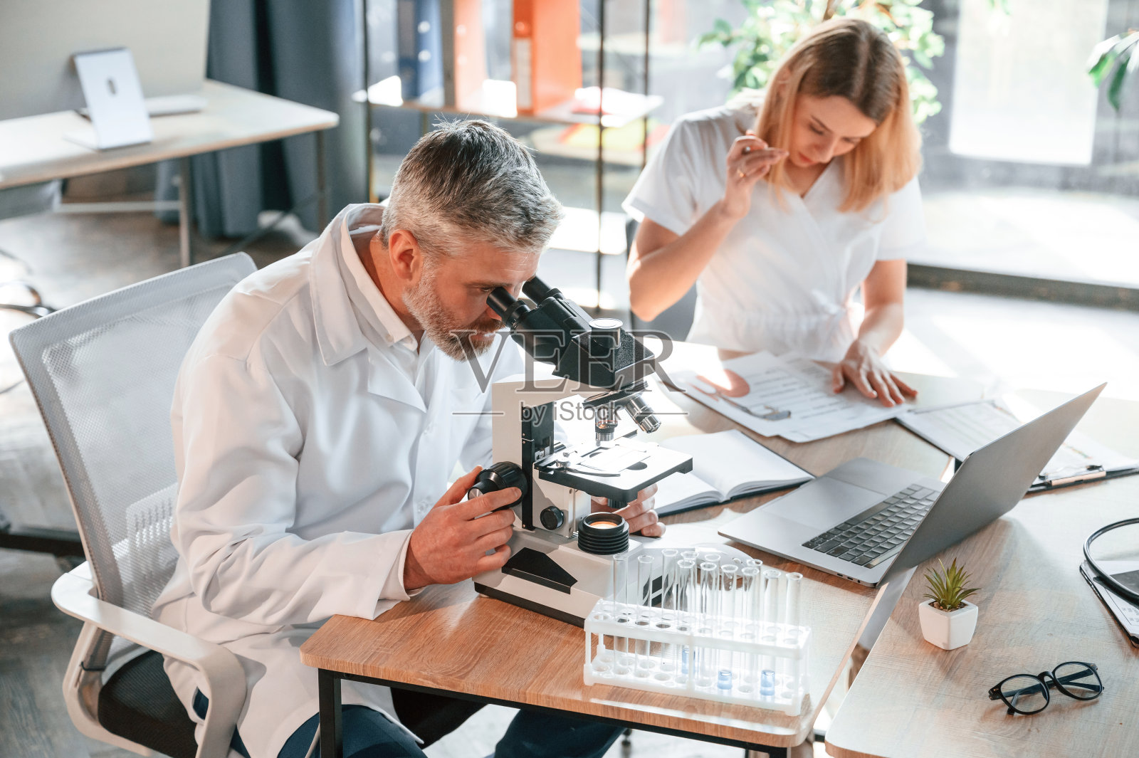 一个人正坐着用显微镜。两个诊所的工作人员一起在室内照片摄影图片