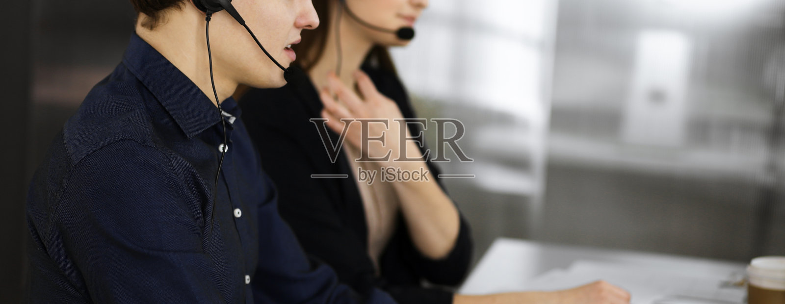 两个戴着耳机的年轻人坐在一间现代化办公室的办公桌前与客户交谈。注意看那个穿蓝衬衫的男人。呼叫中心接线员在工作照片摄影图片