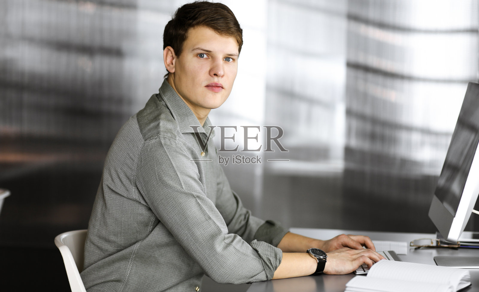 年轻的成功的商人和程序员在绿色衬衫看着相机,而他正在他的电脑工作,坐在桌子上的一个柜子。办公室里的大头照或商务照照片摄影图片