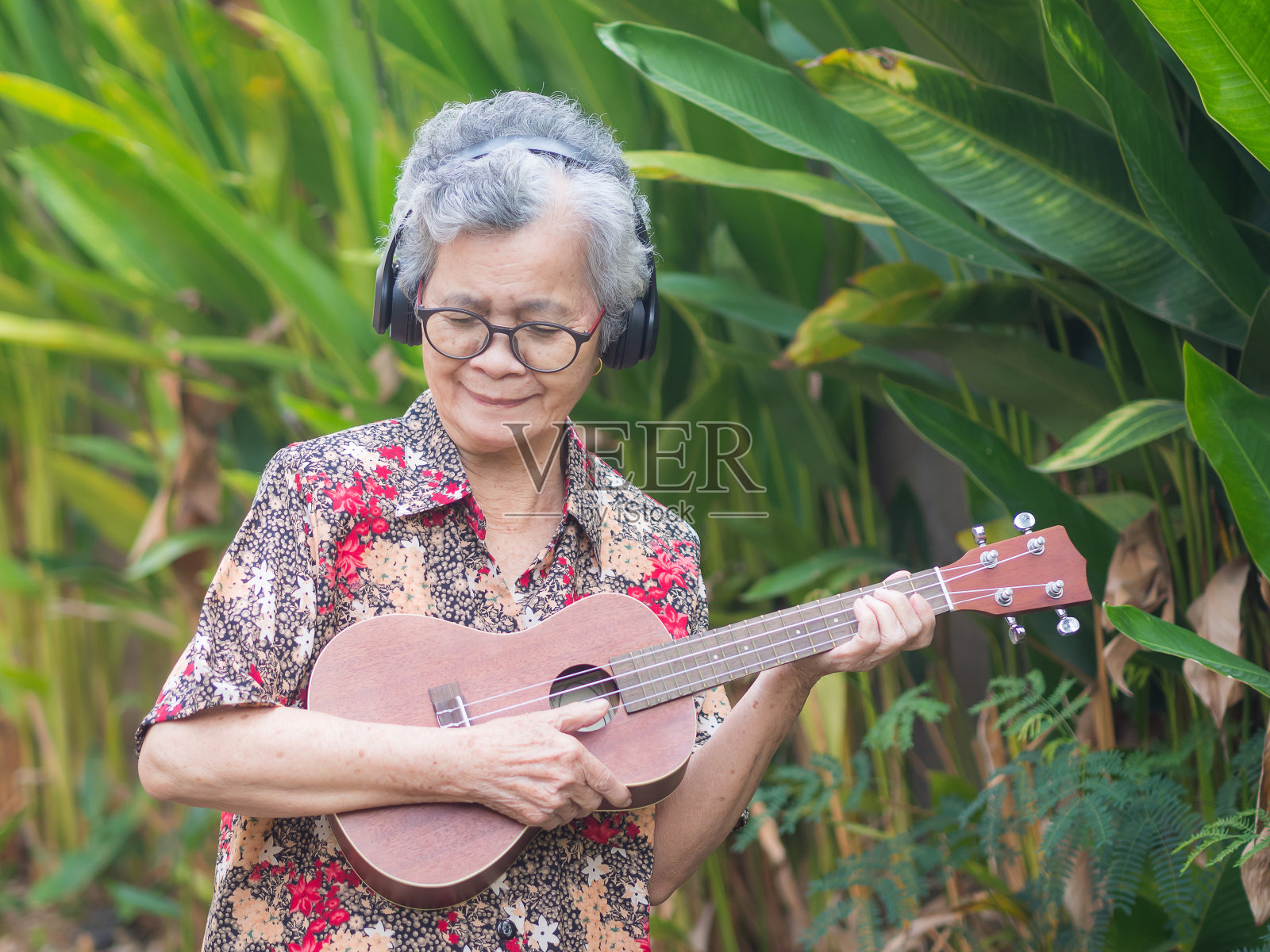 欢快的亚洲老妇人戴着无线耳机,微笑着站在花园里。照片摄影图片