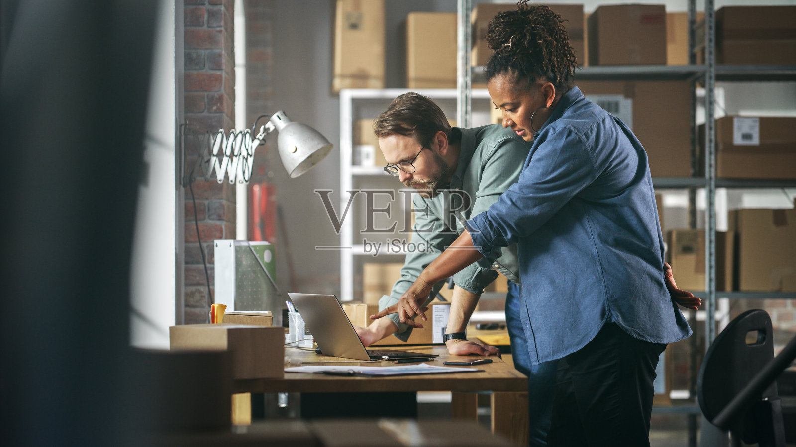 两名员工在他们的网上商店准备网上销售订单。在储藏室工作的男人和女人。非洲妇女使用笔记本电脑,男子确认盒子上的联系方式。照片摄影图片