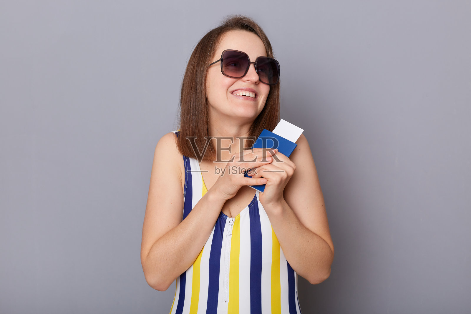 满意的高兴的白人妇女穿着泳衣和太阳镜拿着护照的肖像,很高兴有一个热带岛屿之旅,孤立在灰色背景上照片摄影图片