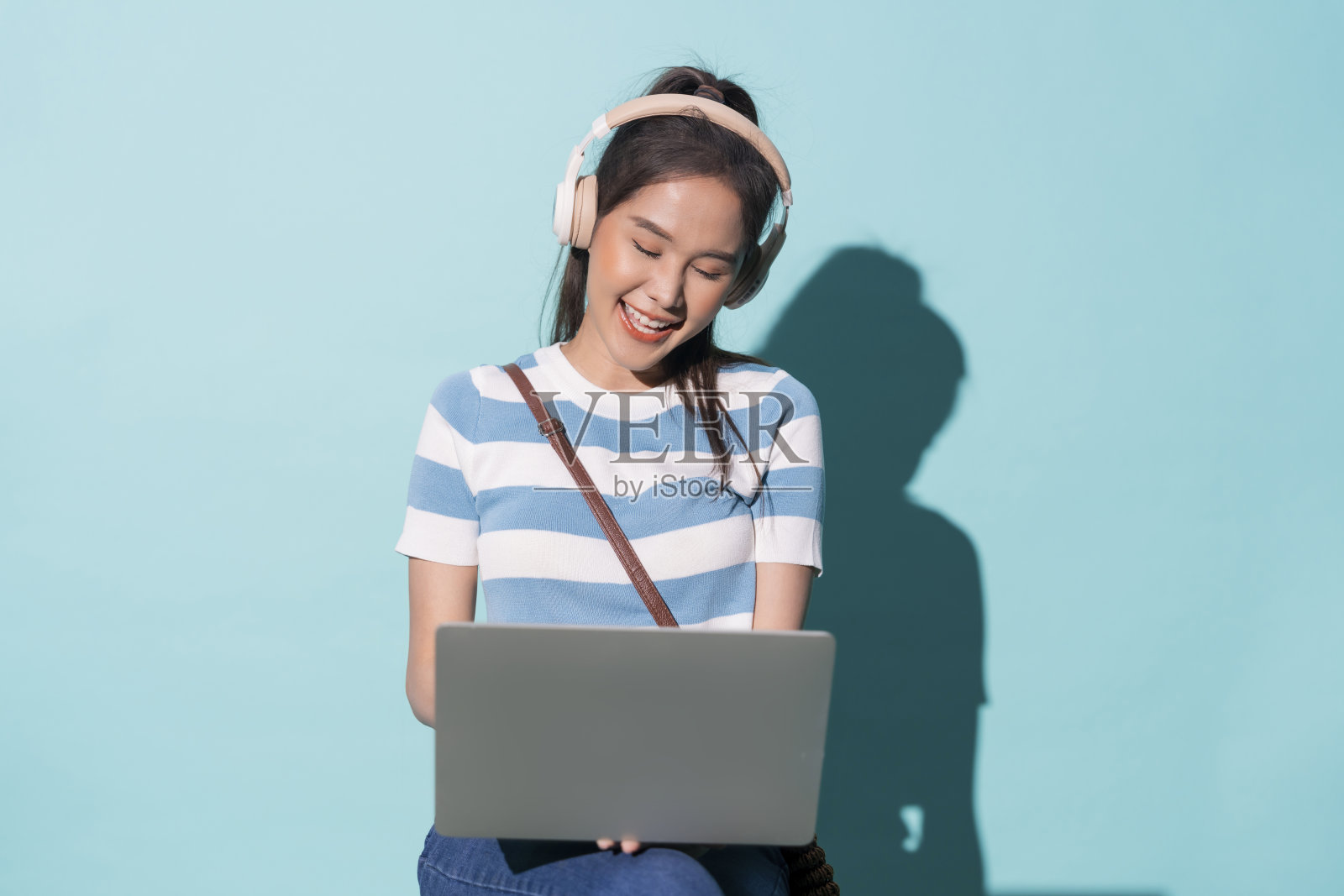 研究大学青少年,亚洲妇女拿着笔记本电脑,戴着耳机,微笑着听着音乐,坐在阳光明媚的白天,自信开朗积极的态度工作室拍摄的彩色背景照片摄影图片