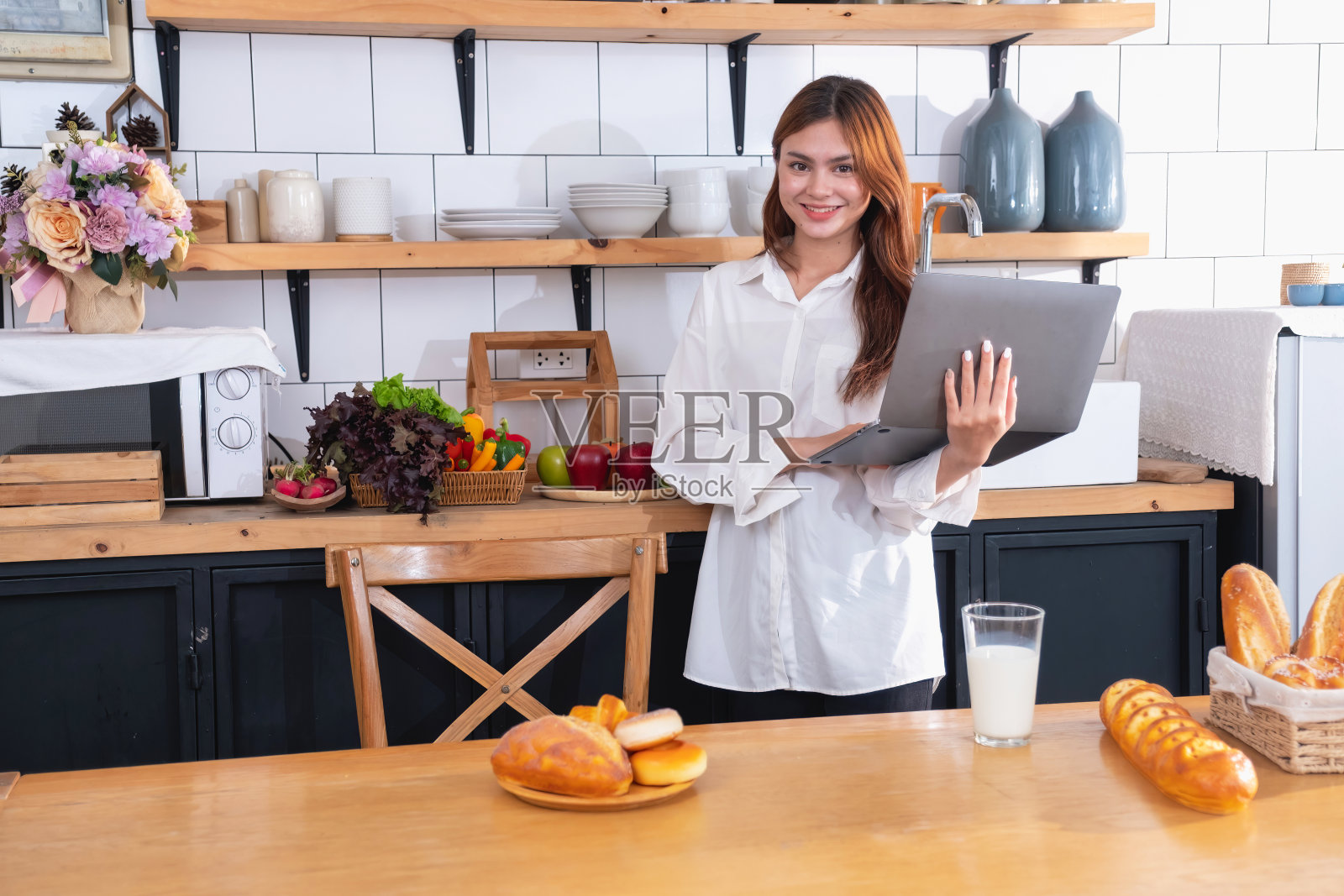一位穿着白衬衫的美女正在厨房里做健康的早餐,有面包、蔬菜、水果和牛奶,并打开笔记本电脑上烹饪课。健康的烹饪理念。照片摄影图片