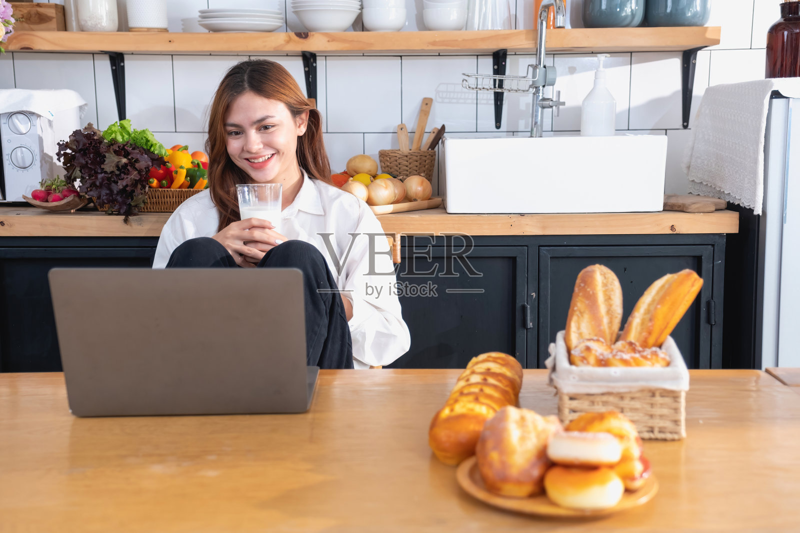 一位穿着白衬衫的美女正在厨房里做健康的早餐,有面包、蔬菜、水果和牛奶,并打开笔记本电脑上烹饪课。健康的烹饪理念。照片摄影图片