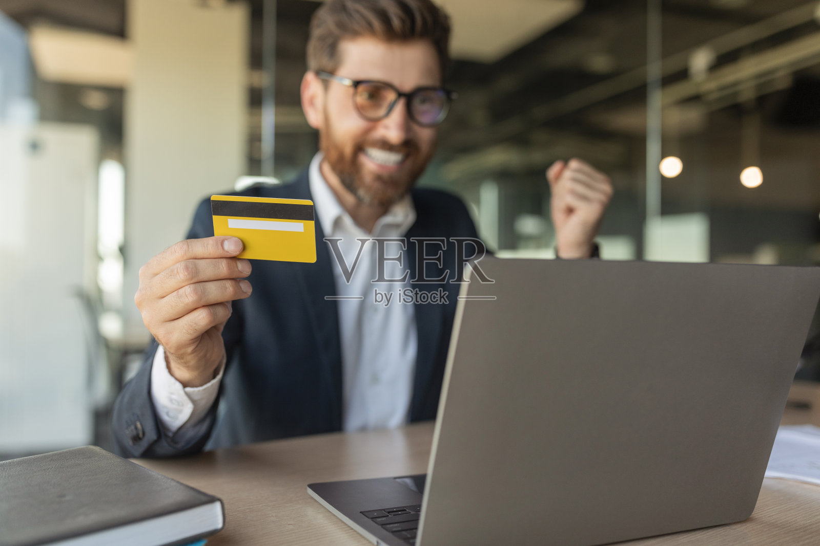 欣喜若狂的商人取得成功的姿态,手持信用卡在笔记本电脑前坐在办公室内部的桌子照片摄影图片