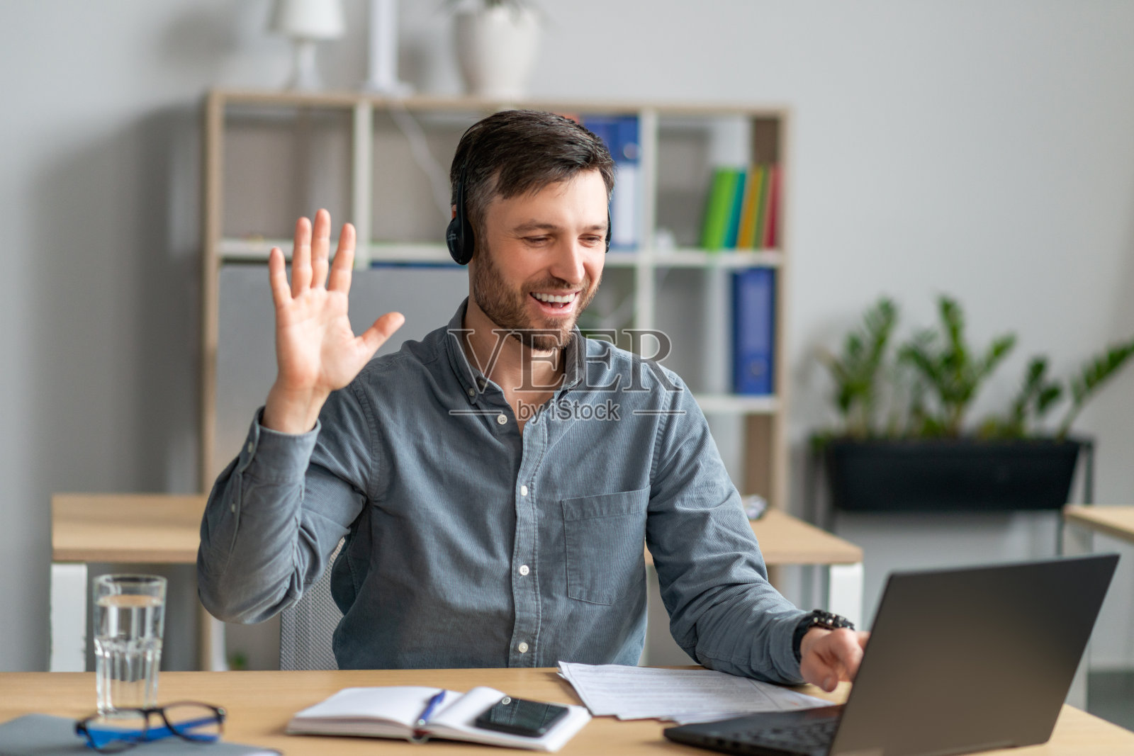 快乐成熟的白人商人戴着耳机在笔记本电脑上工作,对着网络摄像头挥手,坐在办公室里照片摄影图片