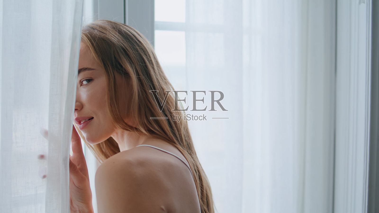 诱人的女人看着镜头从窗帘近。调情女孩独自摆姿势照片摄影图片