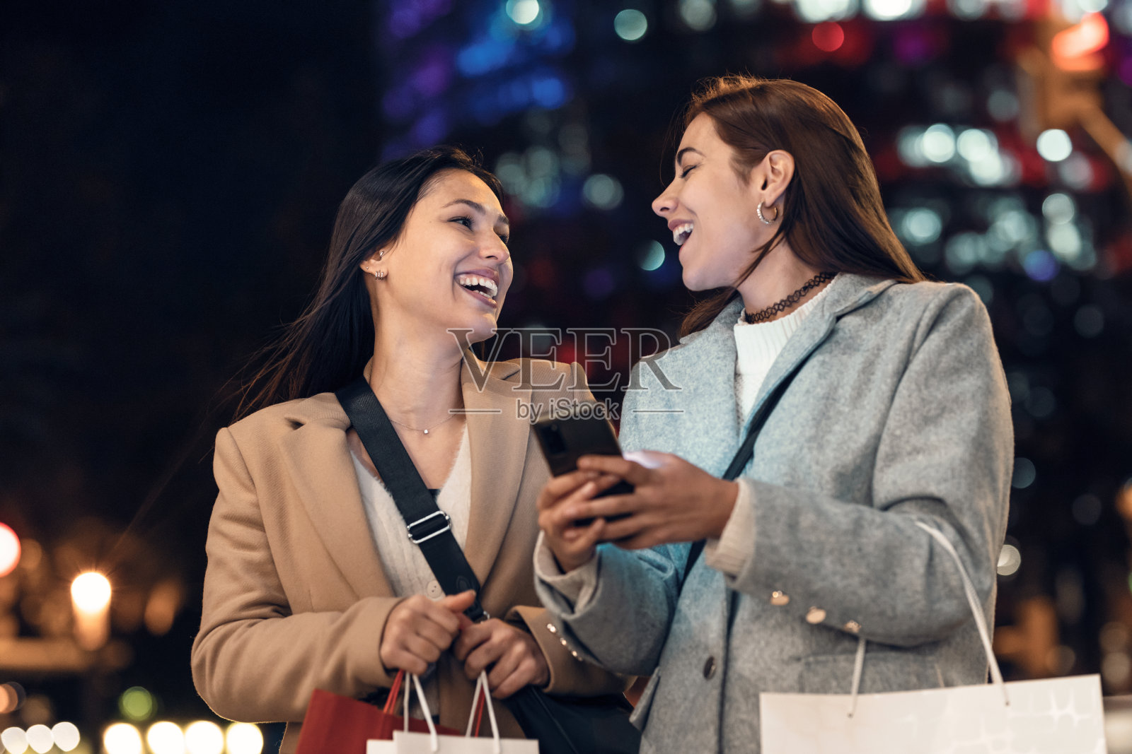 两个美丽的朋友一边聊天,一边用智能手机,背着购物袋走在城市的街道上。照片摄影图片