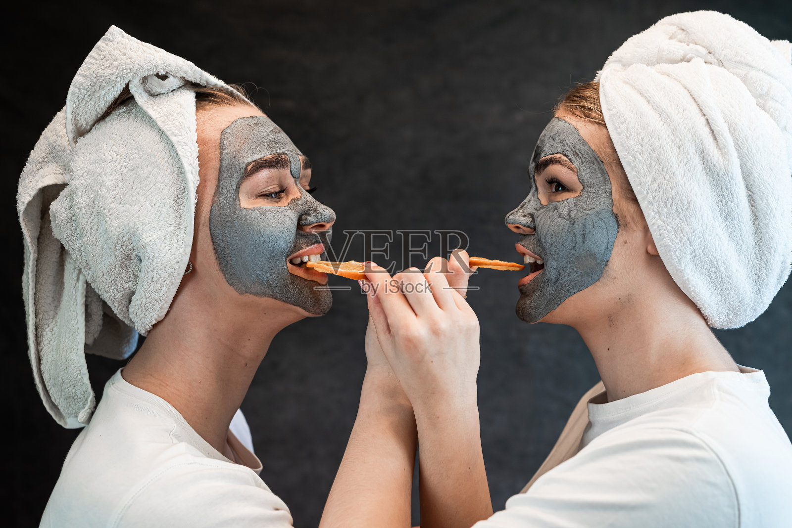 两个白人妇女用白色毛巾敷泥面膜,把橙色隔离在黑色上照片摄影图片