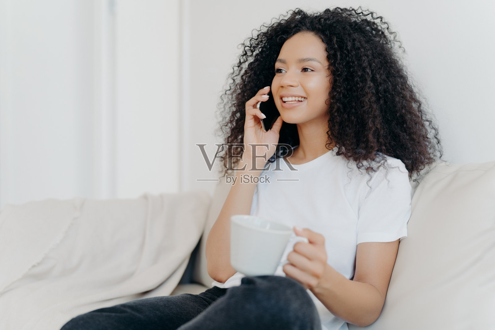 正族黑发女子侧身拍照打电话,喝着热饮,端着白色杯子,坐在客厅舒适的沙发上,听到喜讯,开心地笑着照片摄影图片