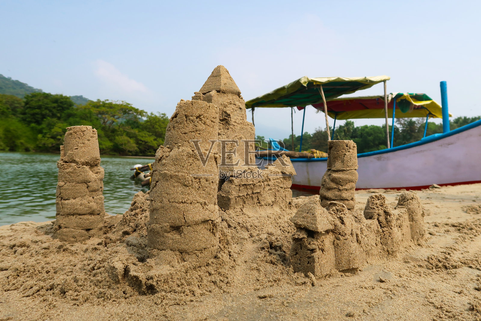在印度果阿的海滩上建造的复杂沙堡堡垒的特写图像,用水桶和铲子制作,并修改为围绕外部,大海和船的墙,背景是平底船照片摄影图片