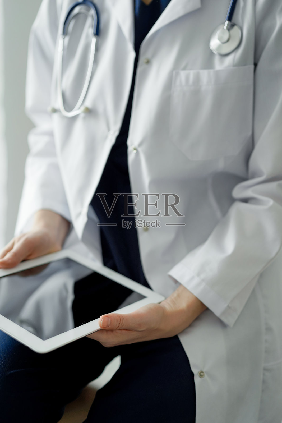 不明女医生站在诊所窗口边使用平板电脑或触摸板。深蓝色的医生工作服。医学的概念照片摄影图片