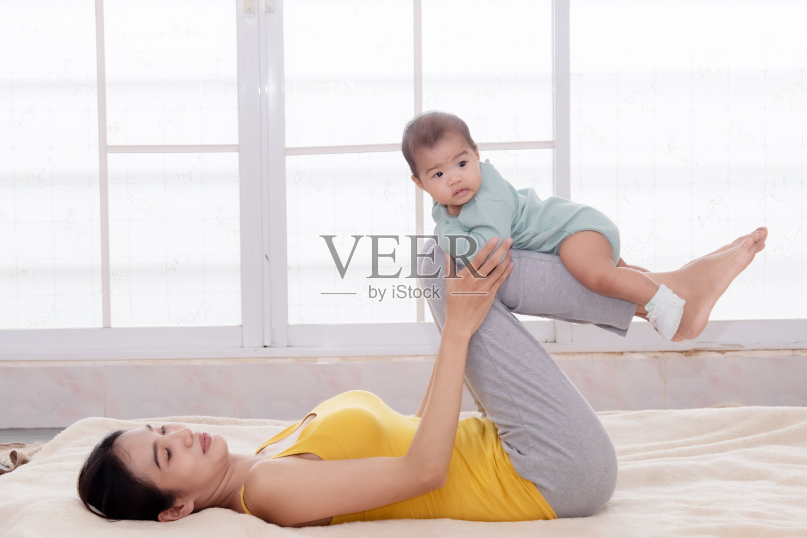 美丽的亚洲母女在家练瑜伽。年轻的妈妈把在床上玩飞机的婴儿抱起来。快乐的母亲在空中抱着婴儿。女人和小孩一起玩,充满爱和关怀。照片摄影图片