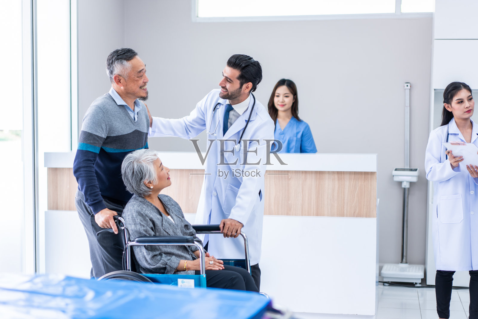 成熟的医生走过走廊去医院看望病人。有魅力的专业男医生正在诊所走廊里为轮椅上的病人做体检。照片摄影图片