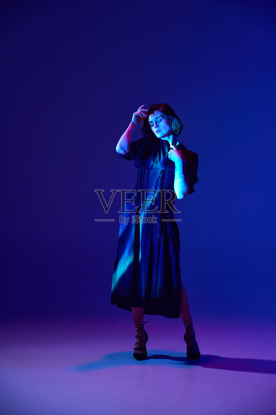 一个迷人的女人穿着黑色及地长裙和高跟鞋,在深蓝色的霓虹灯背景下,在镜头前摆出平静的表情照片摄影图片
