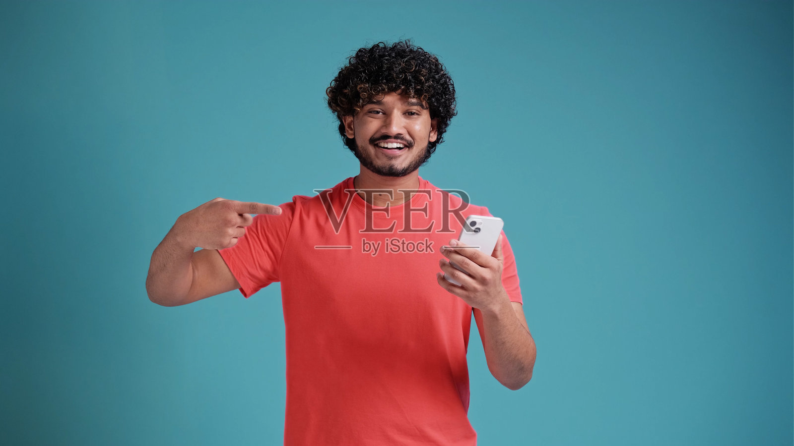 20多岁的印度大胡子年轻男子,喜气洋洋、兴高采烈,手里拿着手机,用食指指着智能手机,身穿珊瑚色t恤,蓝色工作室背景照片摄影图片