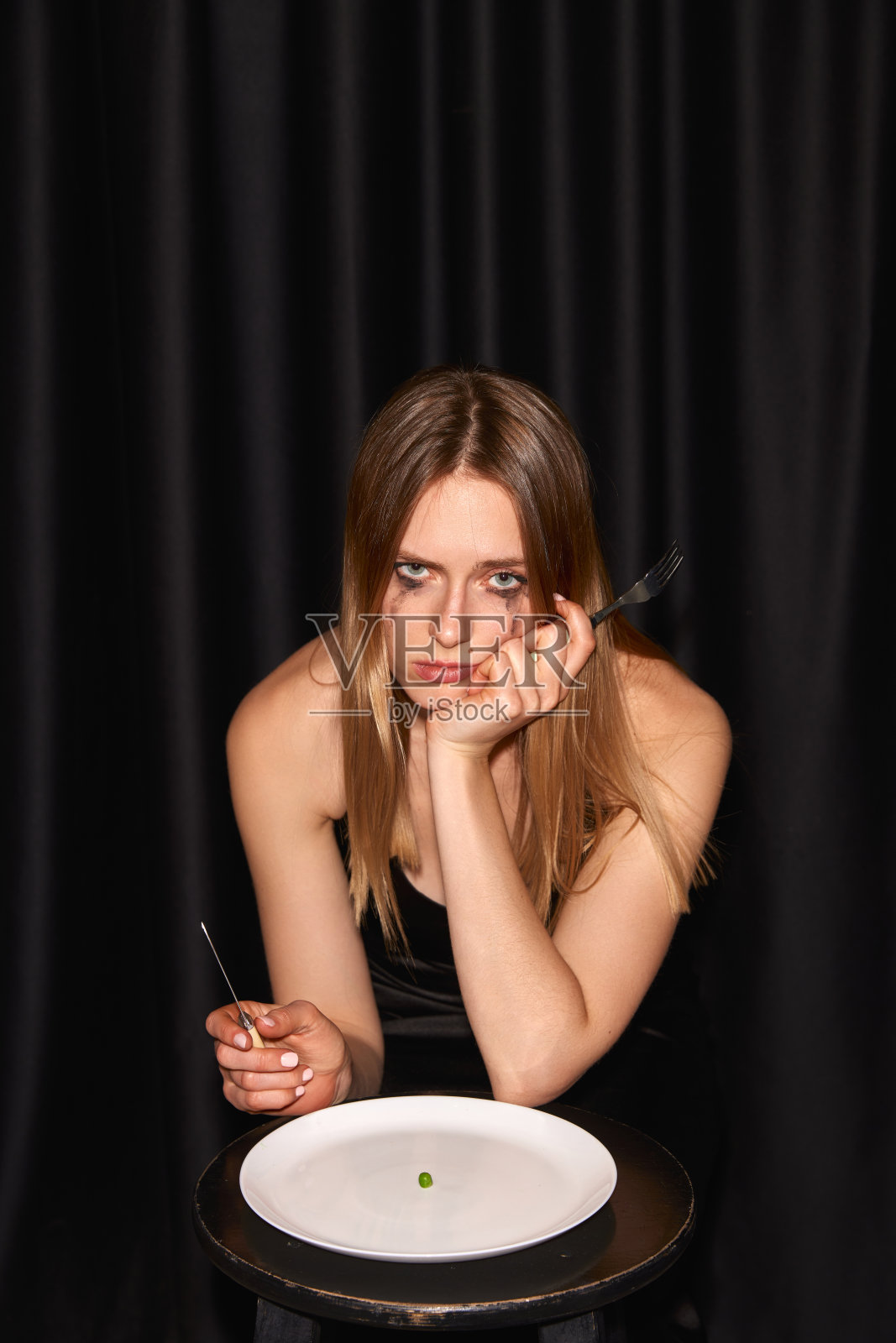 一个年轻苗条的女孩,时尚模特,坐在桌子旁,开始吃几乎空盘子的黑色背景照片摄影图片