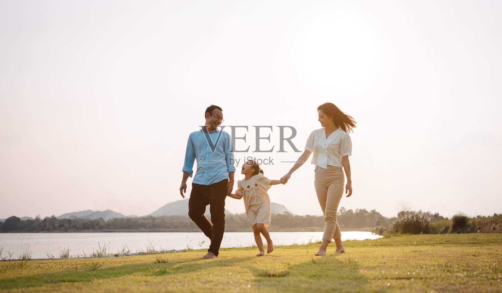 幸福的家庭在公园夕阳下。周末一家人在河边的草地上一起跑步,父母牵着孩子的手。健康人寿保险计划概念。照片摄影图片