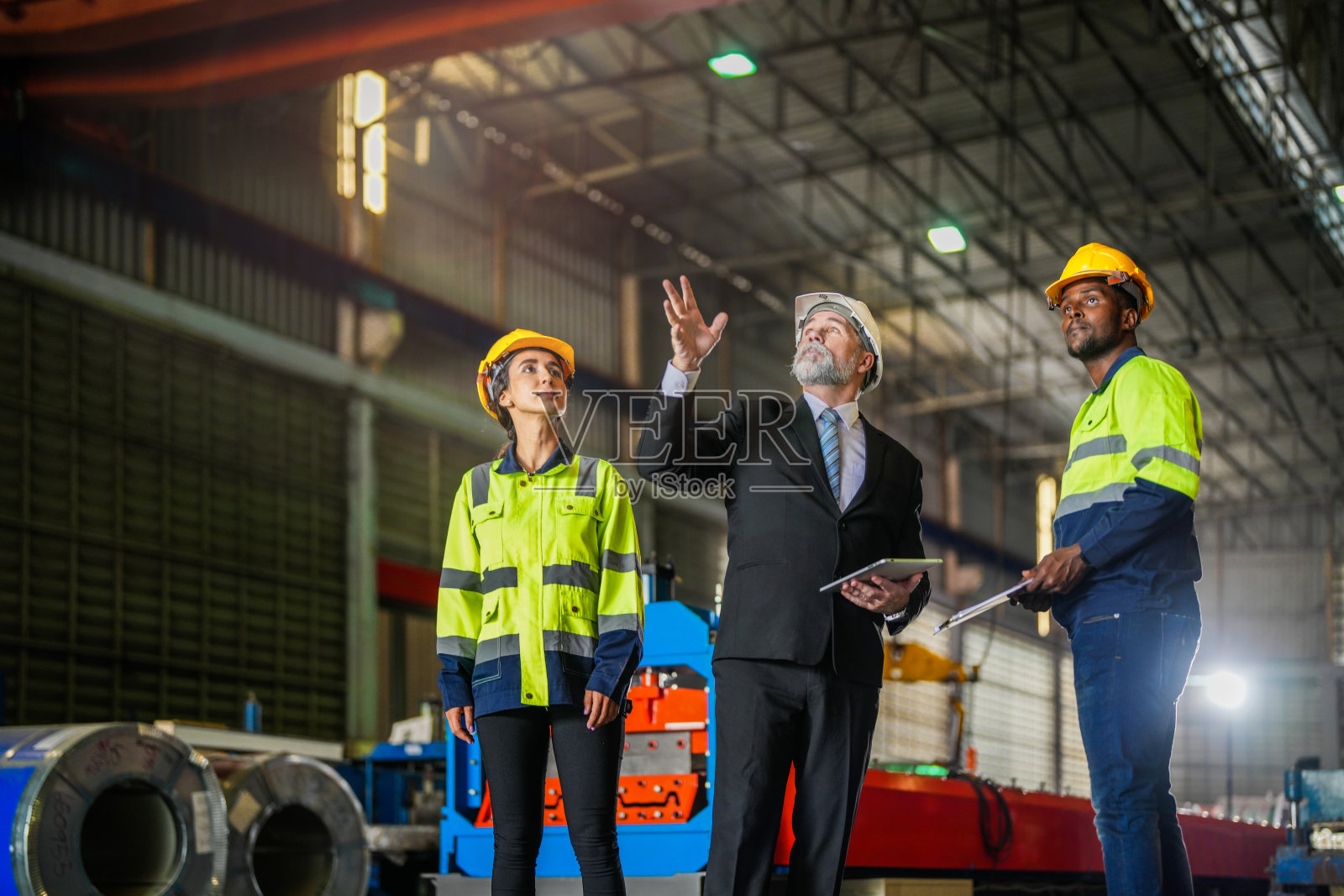 老年经理与员工工程师女和非洲男一起视察。小组检查厂房建筑结构。仓库用钢屋面结构和机器安装。照片摄影图片