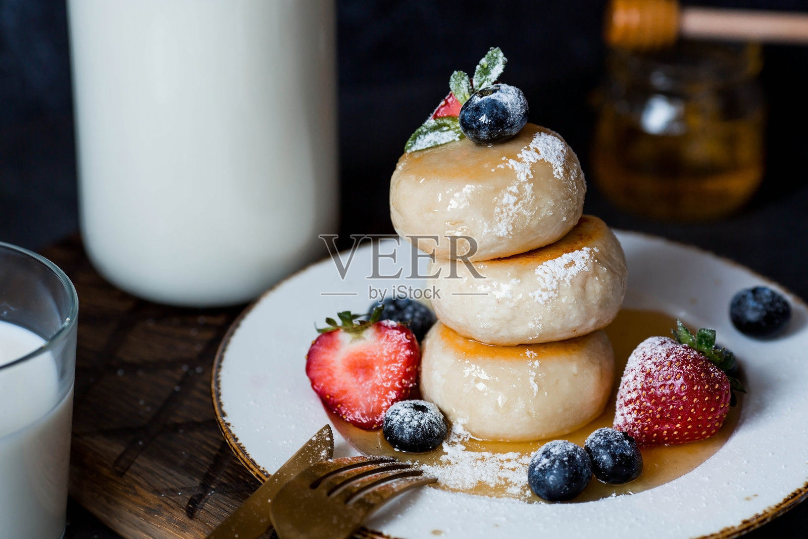 农舍奶酪煎饼,俄罗斯奶酪蛋糕,新鲜蓝莓,草莓,蜂蜜和薄荷叶放在一个时尚的深色背景的木板上。自然产品。健康美味的早餐。照片摄影图片