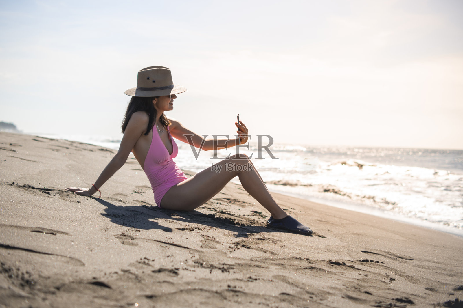 一名年轻的成年女子坐在沙滩上逆光用智能手机打视频电话照片摄影图片