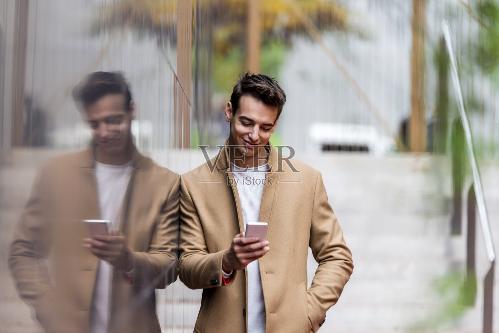 时尚帅气的年轻人穿着外套站在户外,靠在墙上使用智能手机照片摄影图片