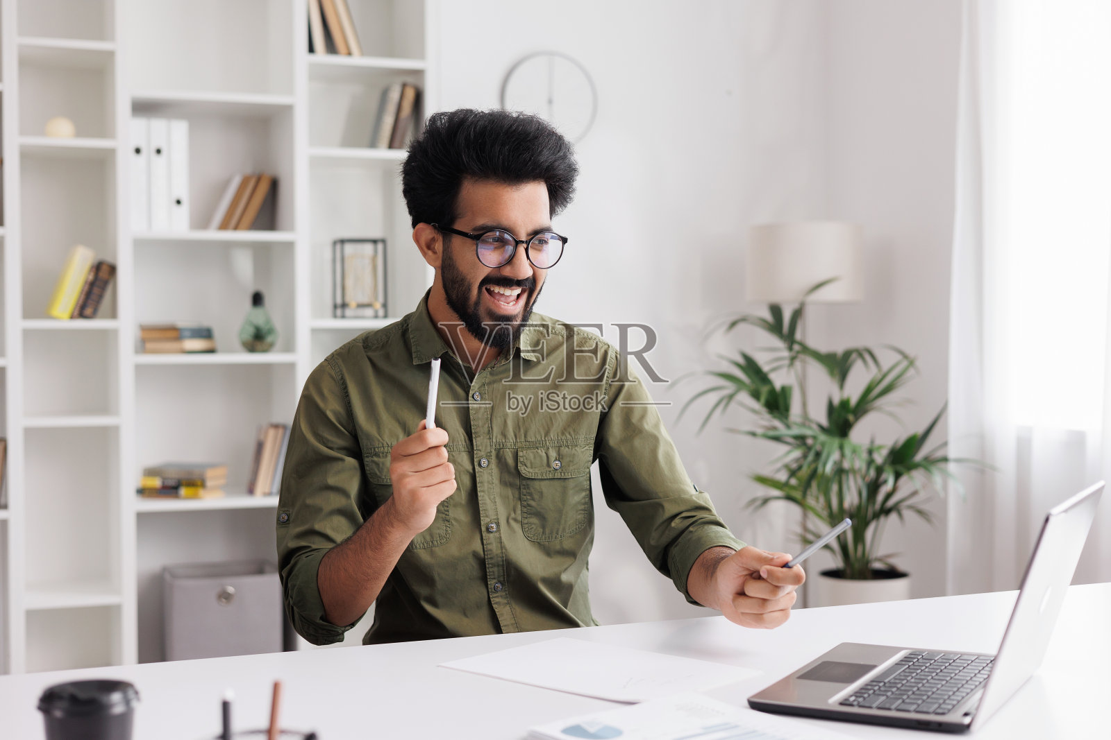 戴着黑色眼镜的印度人满意地查看他的电子邮件,听到好消息很高兴。一个年轻的获胜者在桌子上跳舞庆祝胜利,手里挥舞着笔。照片摄影图片