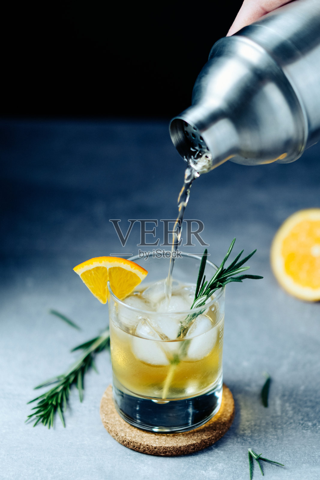 将酒精鸡尾酒从摇酒器倒入玻璃杯中,在木桌上放上橙片迷迭香和冰块照片摄影图片