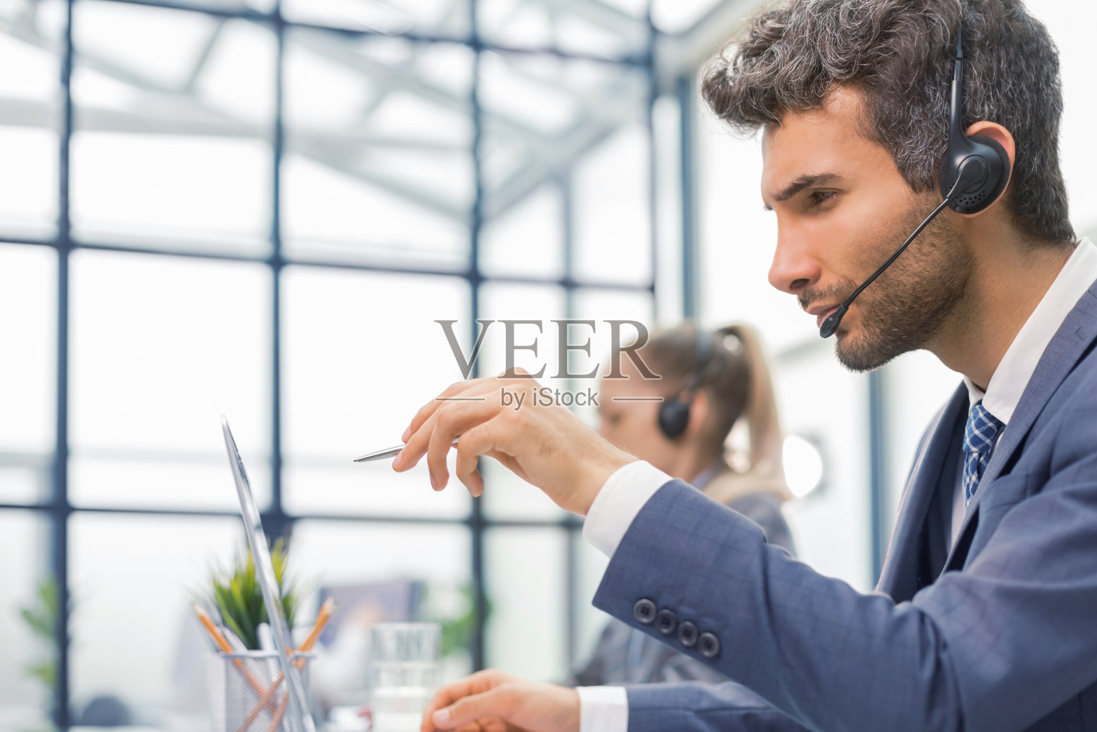 戴着耳机微笑着的男性呼叫中心接线员坐在现代化的办公室里,与同事们一起在网上咨询。照片摄影图片