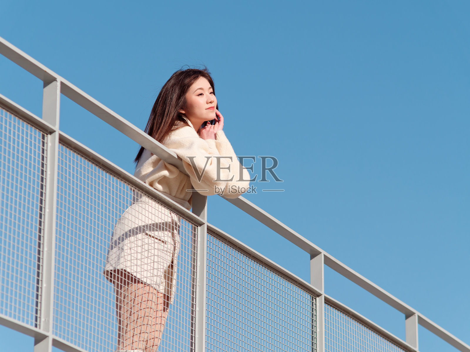 美丽的年轻女子,黑色长发,白色裙子,在阳光明媚的日子里,蓝天背景的桥上摆姿势。情感、人、美、旅行、生活理念。照片摄影图片