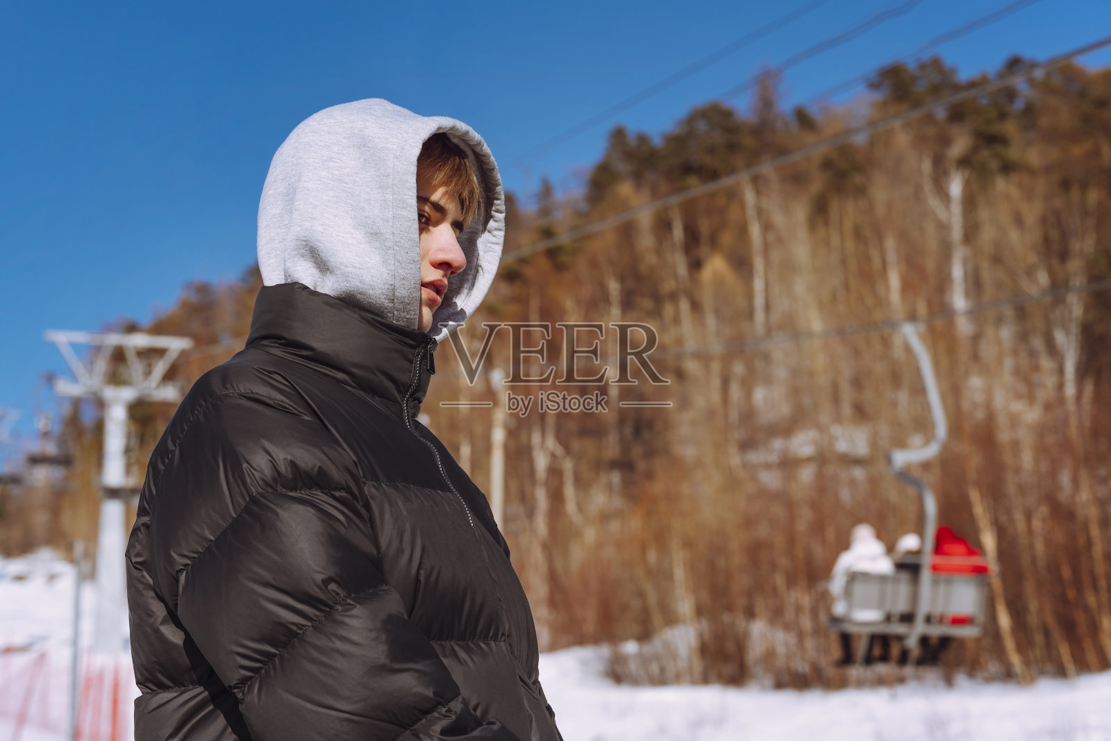 在滑雪胜地,戴着兜帽的少年抬头看着滑雪缆车。照片摄影图片
