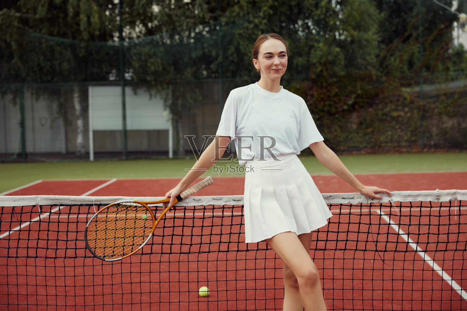 穿着白色衣服的女孩拿着网球拍在球场的网网前摆姿势照片摄影图片