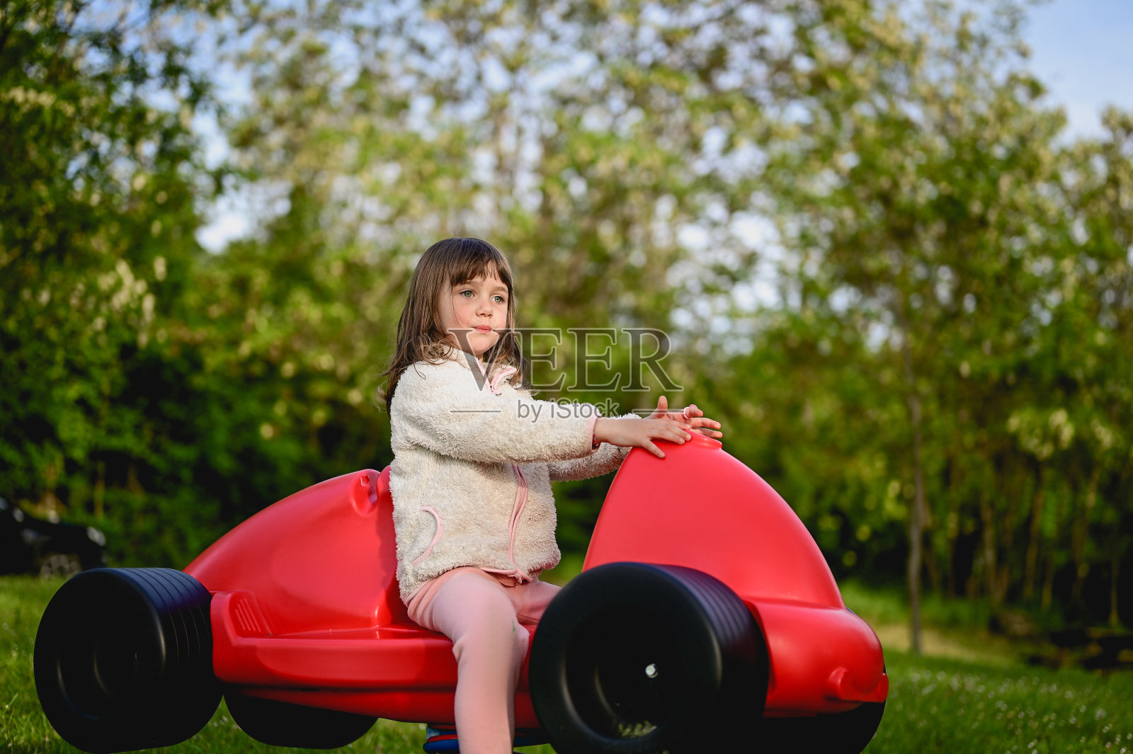 在户外操场上,一个学龄前女孩坐在一辆红色的汽车里。照片摄影图片