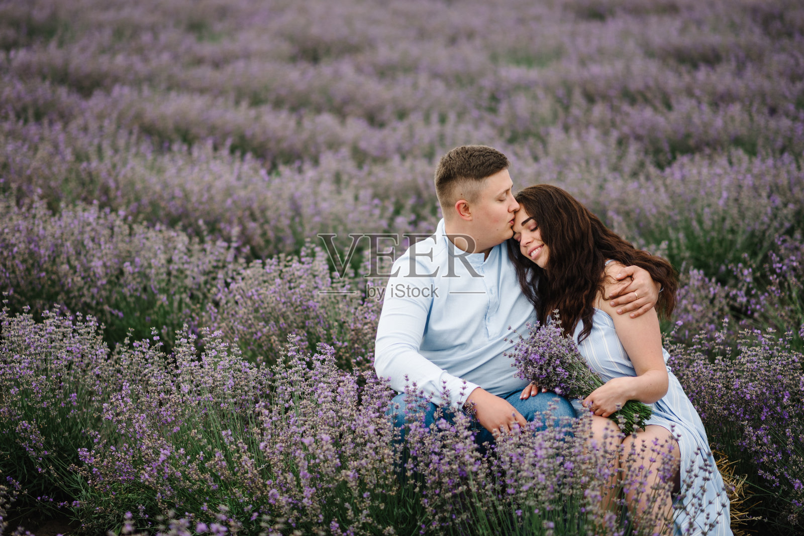 一对恩爱的情侣在日落时分捧着一束薰衣草花亲吻。夫妇坐着享受花香的林间空地,夏日的自然。法国,普罗旺斯。家人在紫色薰衣草地里拥抱。蜜月旅行。照片摄影图片