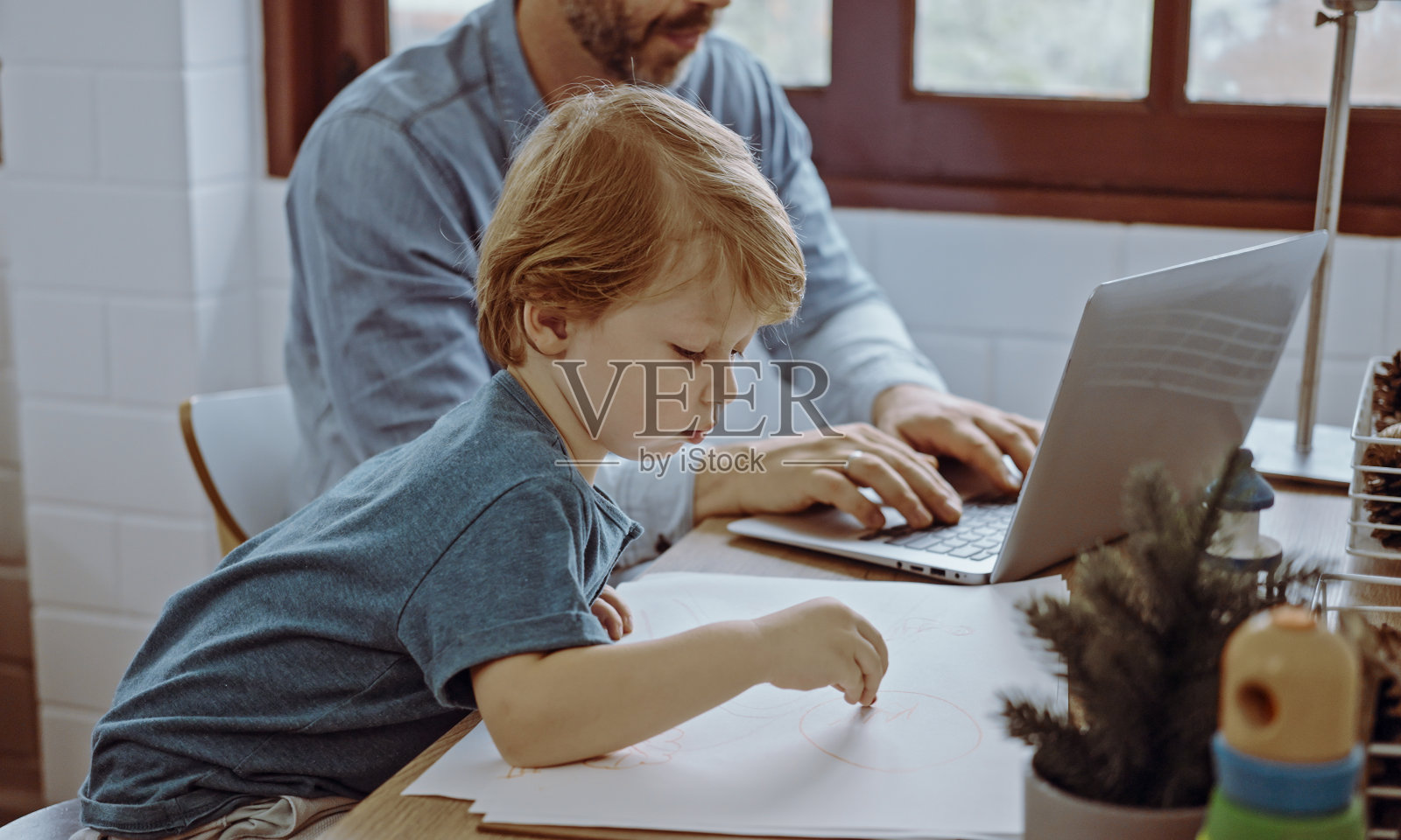 父亲在家办公,用笔记本电脑坐在桌子旁和可爱的小儿子玩耍,几乎打扰。照片摄影图片