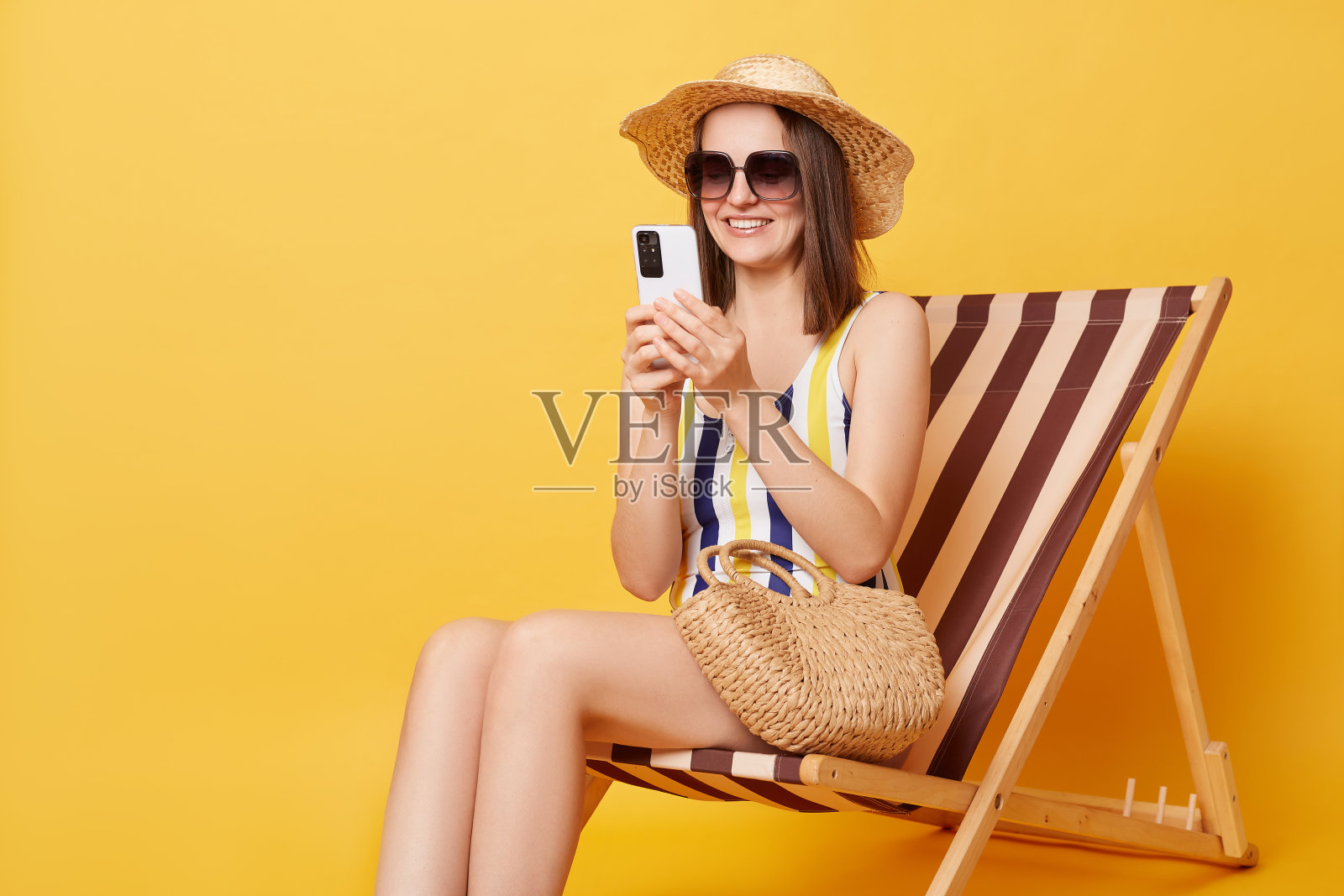 快乐积极的女人用智能手机在沙滩上度过暑假,戴着太阳镜,穿着泳衣,微笑着上网查看社交网络。照片摄影图片