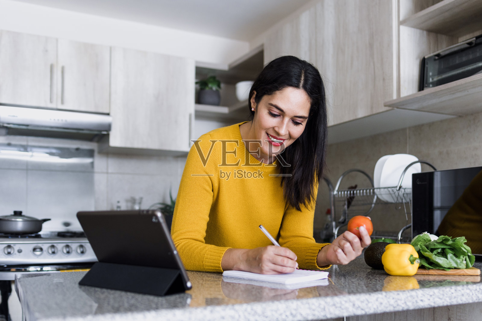 拉丁美洲,西班牙裔女性,在墨西哥家中的厨房里做饭并在笔记本上写菜谱照片摄影图片