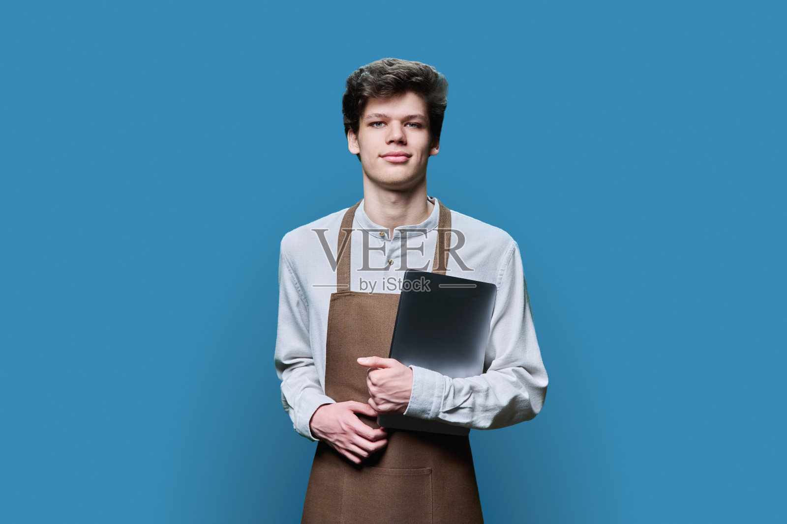 系着围裙拿着笔记本电脑的年轻男子,蓝色背景照片摄影图片