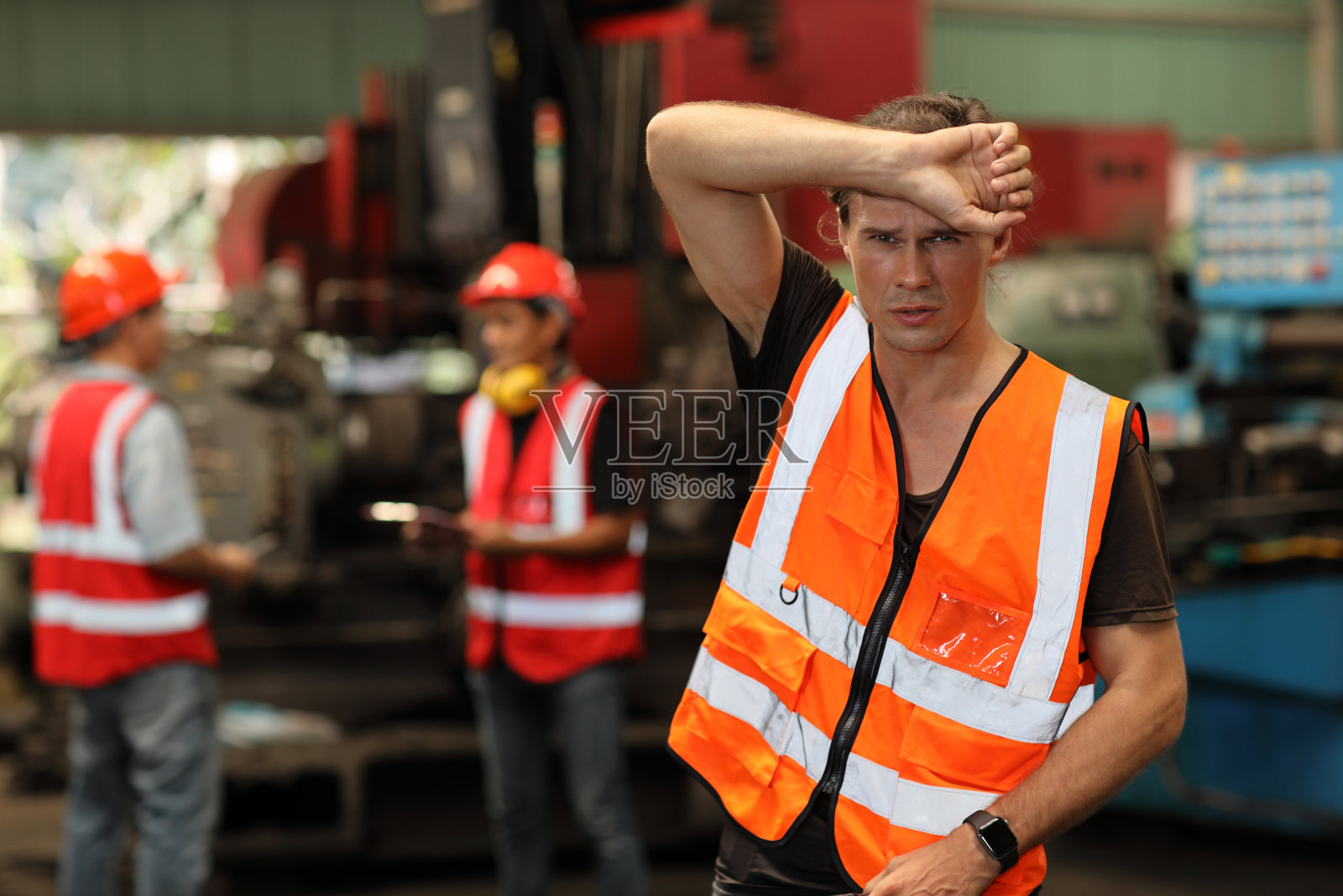 在重工业制造厂,疲惫的技师穿着防护服站着休息,戴着安全帽在检查完工业机器过程后擦去汗水照片摄影图片