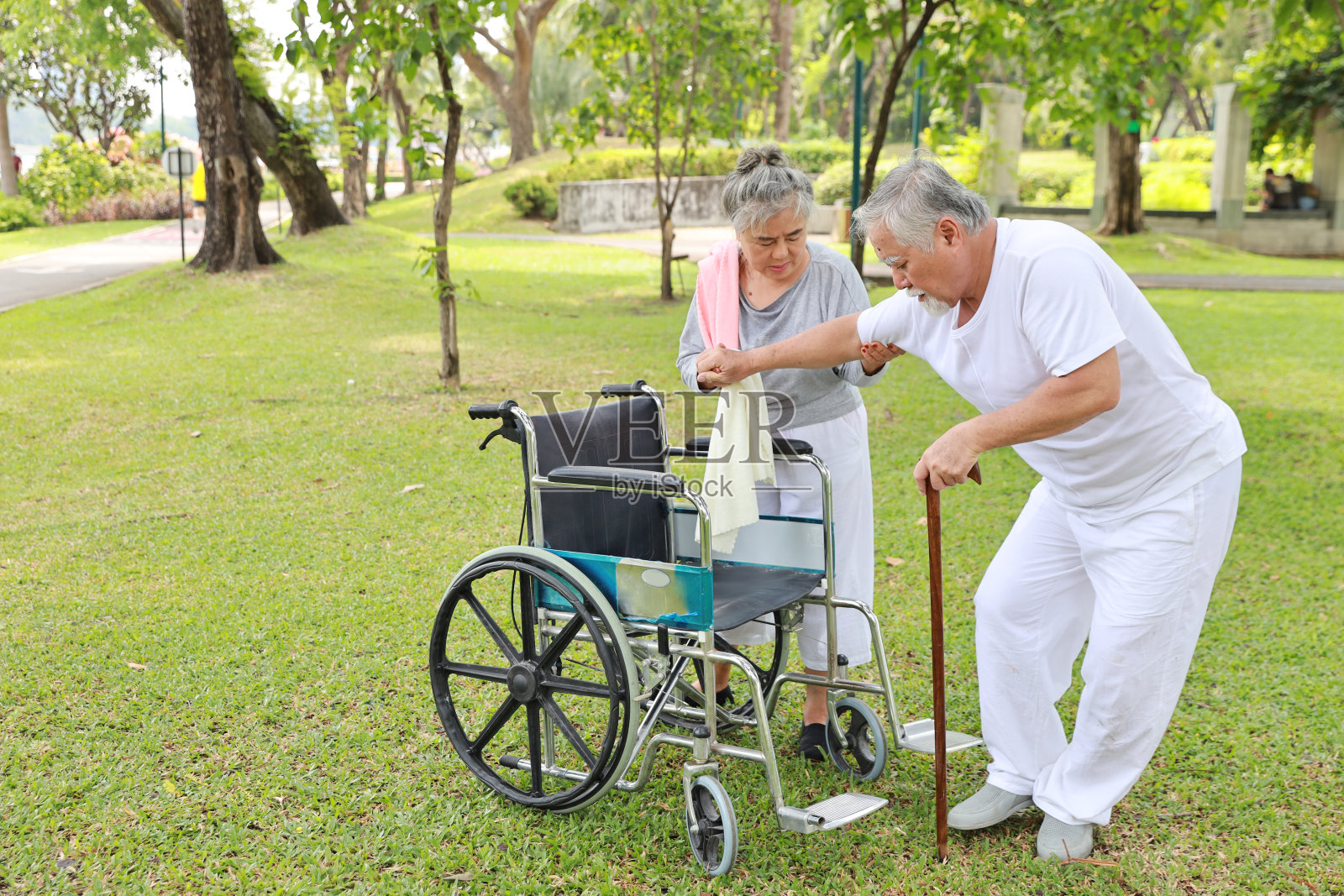 亚洲老年妇女或护理员在公园户外帮助老年男子用轮椅行走。年老的妻子在养老院外照顾和支持年老的丈夫。照片摄影图片