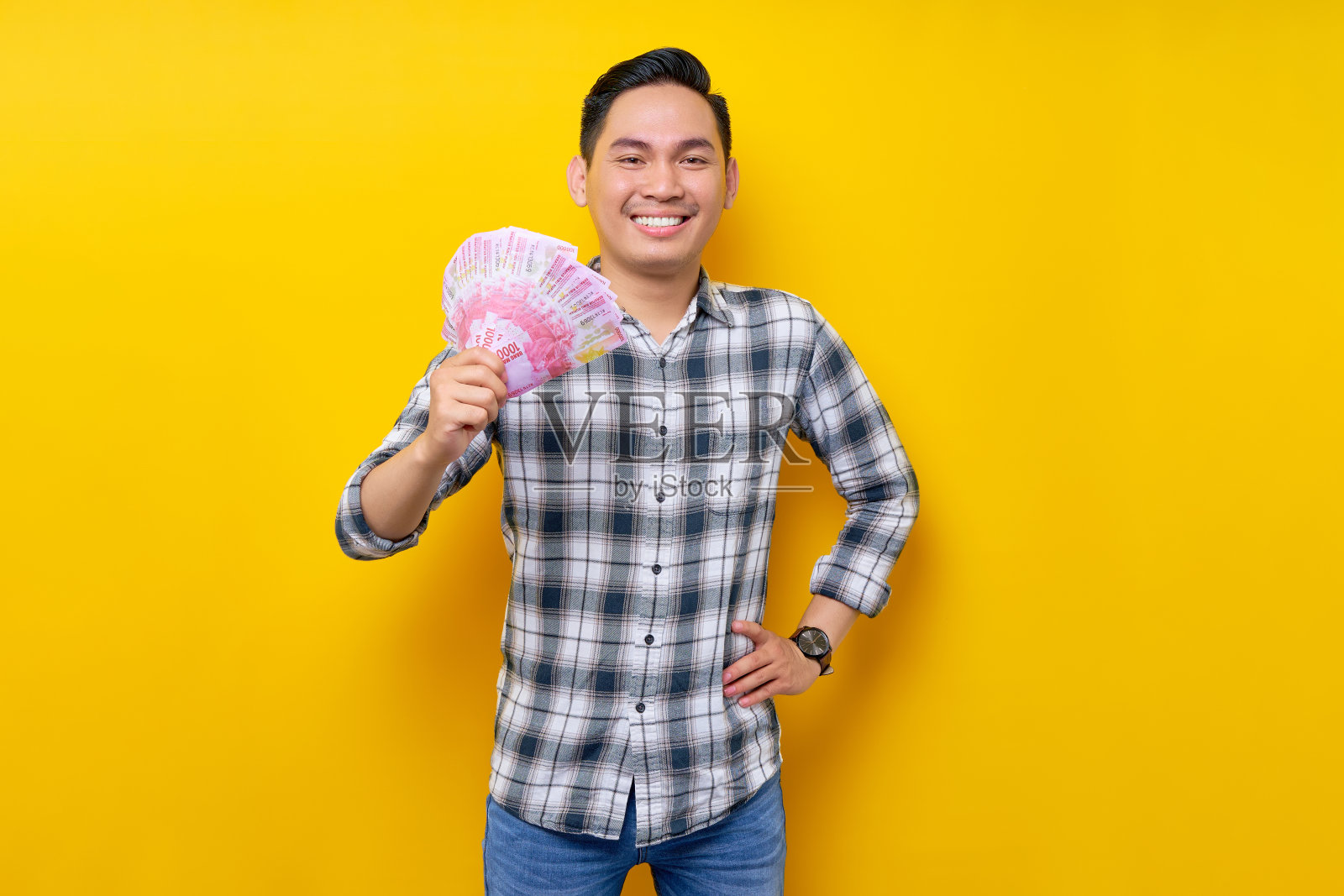 穿着格子衬衫微笑的年轻亚洲男子站着摆姿势,手里拿着印尼盾纸币,黄色背景上有一个自信的表情。人们的生活方式理念照片摄影图片