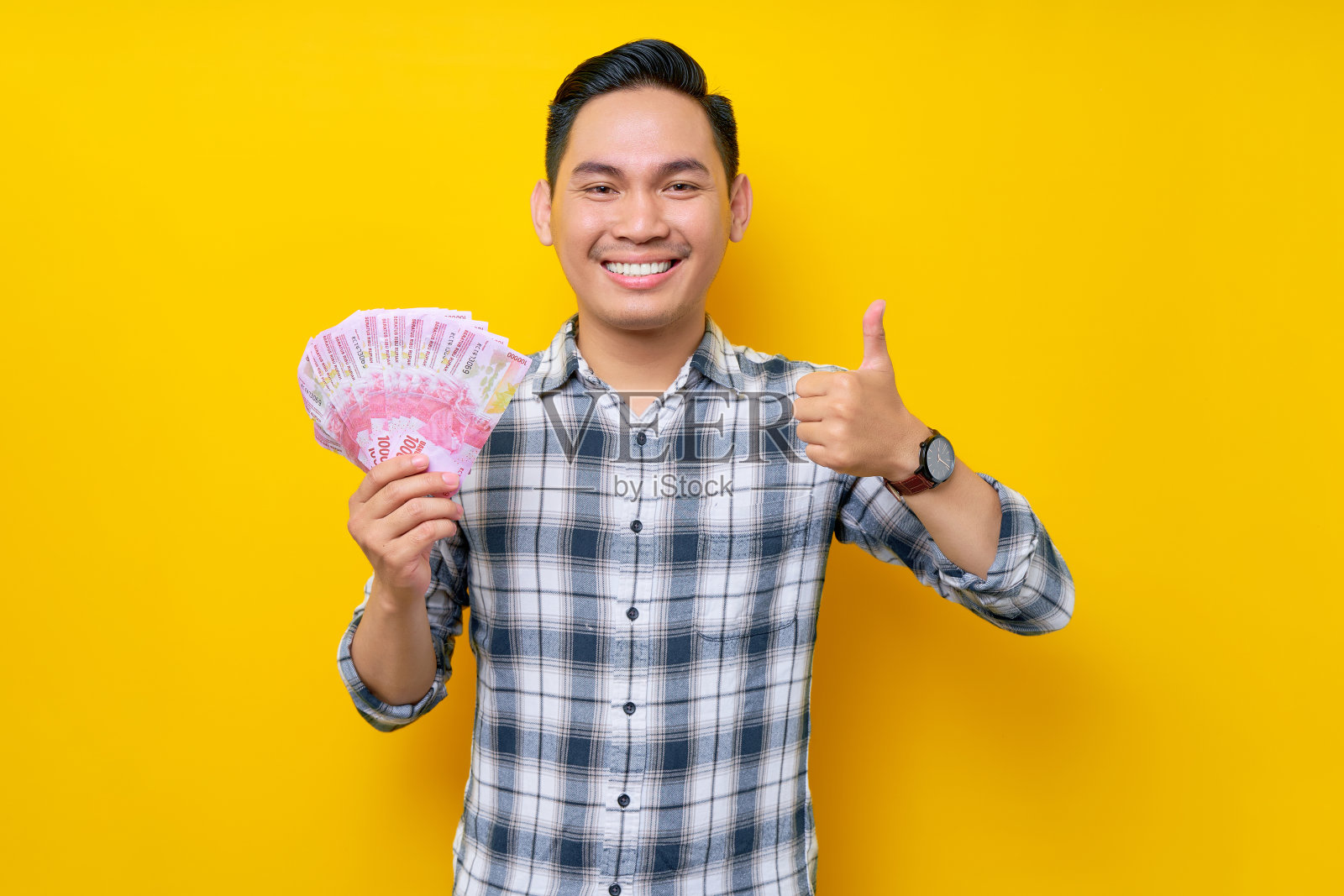 穿着格子衬衫微笑的年轻亚洲男子的肖像,手持一扇印尼盾钞票,并在黄色背景上竖起大拇指。人们的生活方式理念照片摄影图片