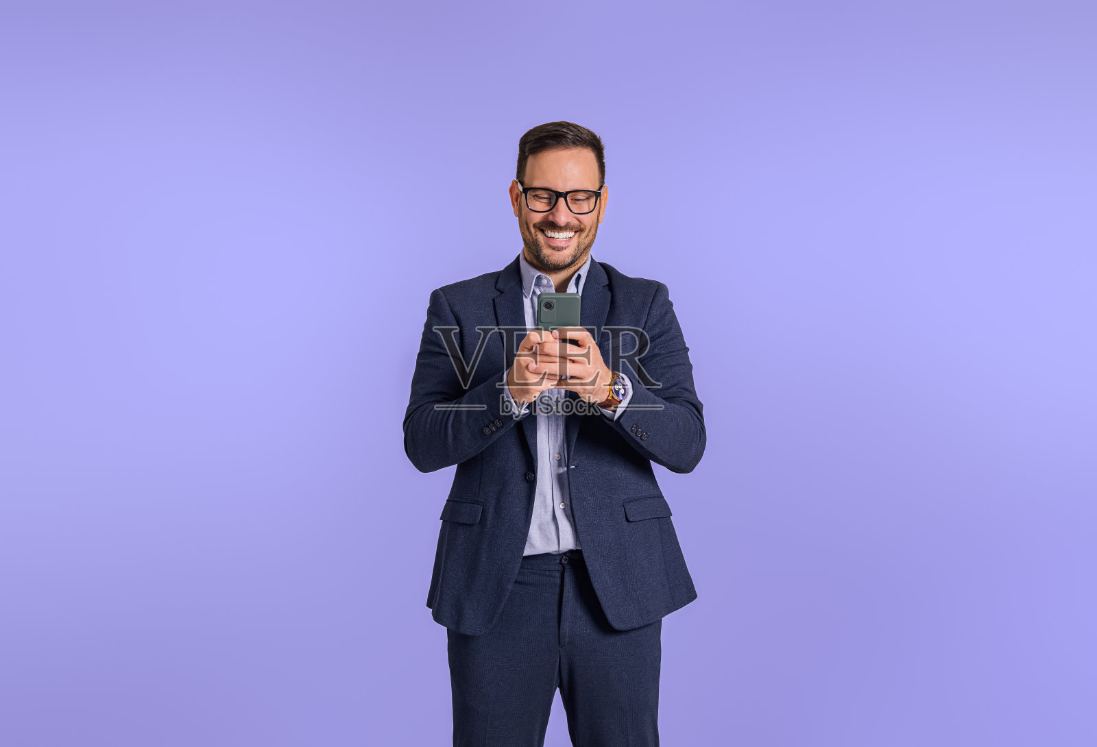 积极的年轻业务经理穿着西装,微笑着通过智能手机远程办公。男性企业家用手机做社交媒体营销,独自站在蓝色背景上照片摄影图片