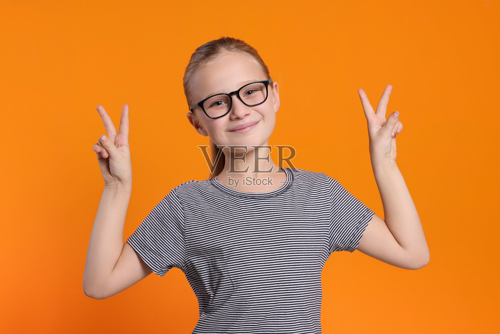 可爱的女孩戴着眼镜,在橙色背景上展示胜利的姿态照片摄影图片